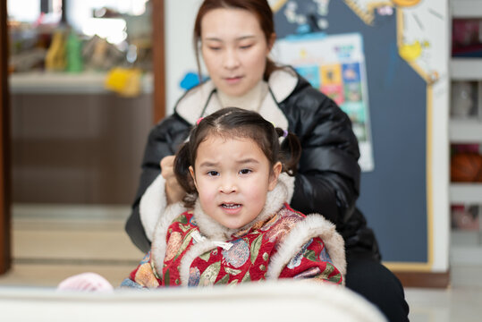 The young Chinese  mother is combing and binding her daughter's hair. Her daughter is wearing traditional Chinese clothes. They're getting ready for celebrating  Chinese New Year.