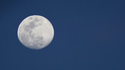 A bright full moon shining in a dark blue night sky surrounded by soft clouds and lunar craters visible