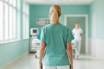 Back view of a nurse in scrubs in a bright hospital hallway with another healthcare professional in the distance.
