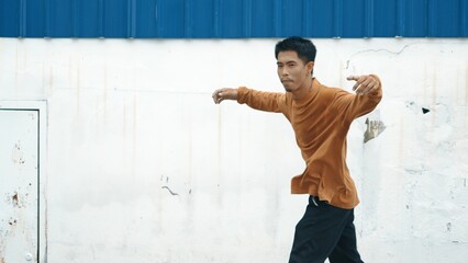 Hispanic man stretch arms and dance break dancing in front of wall. Motion shot of stylish street dancer in casual outfit practicing dancing in hip hop style. Outdoor sport 2023. Background. Hiphop.