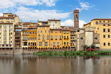 Fototapeta premium Colorful medieval houses sit above the Arno River in Florence, showcasing vibrant facades and historical architecture under blue cloudy sky.