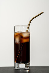 Glass of cola beverage with ice cubes with a long metal golden straw, standing on a black coaster,...
