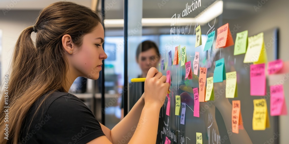 Poster a focused young woman writes on a glass board covered in colorful sticky notes, discussing planning 