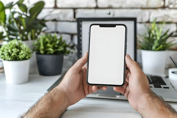 Hands holding smartphone with blank screen, laptop and plants in background.