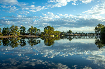 Obraz premium Wimmera River. Riverbank with water reflection of beautiful cloudy sky, residential houses and Anzac Centenary Bridge in the distance. Horsham, VIC Australia.