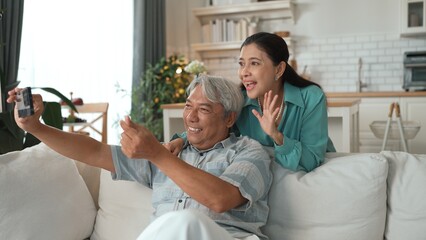 Happy grandfather and grandmother making video call with their son at home. Elder man holding smart phone and sitting at sofa while old woman greeting with cheerful vibe at modern house. Myrmidon.