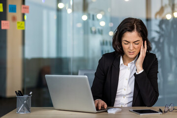 Fototapeta premium Businesswoman in office suit using laptop, experiencing discomfort and stress. Concept of workplace challenges and focus. Office supplies, including phone, visible on desk.