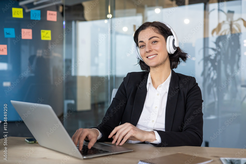 Poster Businesswoman in office wearing headphones working on laptop, smiling at desk. Engaged in virtual meeting, focused on work, technology, communication, productivity. Modern and confident atmosphere.