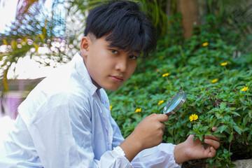 Asian male student goes into the field to study plants, using a magnifying glass to look at flowers and various plant components from the plants.