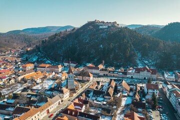 Vibrant rooftops of Rasnov stretch beneath the shadow of Piatra Craiului, where the sun illuminates...