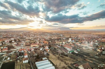 Fototapeta premium As the sun sets behind the mountains, an aerial vista reveals the charming town of Rasnov, nestled among fields and dotted with historical architecture under dramatic skies in Brasov, Romania