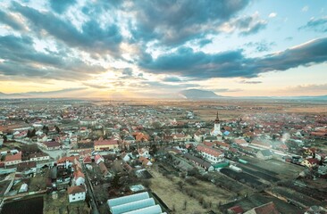 Fototapeta premium Majestic mountains loom in the background as quaint rooftops hug the valleys of Rasnov and Plaiul Foii. The warm hues of sunset bathe the landscape in an enchanting glow, showcasing nature's beauty