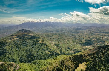 Soaring above the stunning Piatra Craiului mountain range, vibrant green forests and rolling hills stretch towards Rasnov, under a blue sky filled with fluffy clouds