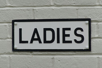 Sign for Ladies toilet. Black lettering on white background on white brick wall.