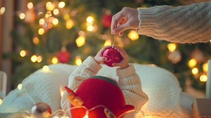 Hand of mother holding red Christmas tree balls with playful baby enjoying near the Christmas tree in holiday, little infant girl in reindeer hat, First Christmas, Christmas and New Year concept.