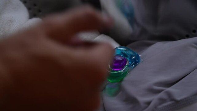 Closeup of colorful laundry detergent pod being placed into washing machine drum, surrounded by white towels and gray clothes, ready for wash cycle. Concept of easy housekeeping domestic life.