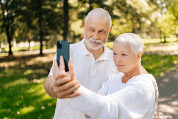 Over shoulder shot of charming elderly couple enjoying video call while strolling in sunny summer park. Joyful gray-haired wife and husband enjoying video call outdoors surrounded by natural beauty.