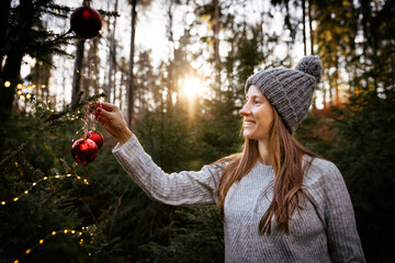 Smiling caucasian woman in woolen sweater hanging a red ball on Chrismas tree in forest. Eco-friendly holiday celebration. Festive new year concept.