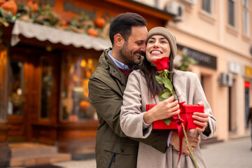 Caucasian man and woman in city setting, man gifting red present and rose to smiling woman. Both wear warm winter attire with woman holding gift and flower.