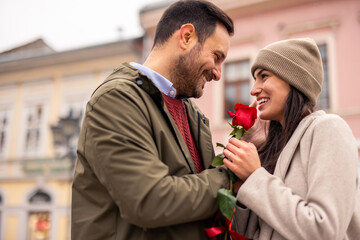 A joyful mid-adult Caucasian couple enjoys a romantic stroll on a city street during Valentine's Day. Dressed warmly, they hold a rose, creating a tender connection.