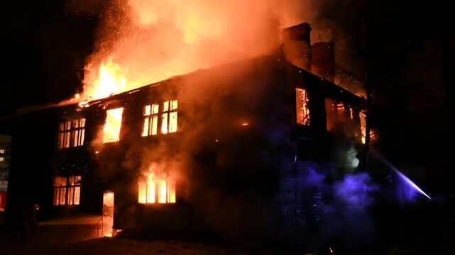 Firefighters are fighting a burning house fire. A fireman is watering the flames in the house at night. The fiery disaster and the work of rescuers. A fire in the Building.