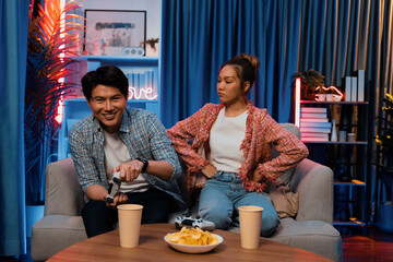 Sitting sofa of happy couple playing video game with joysticks, smiling man playing challenge, raising fist up to be winner the challenge at modern neon blub light living room at home. Infobahn.