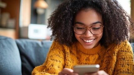 Young african american woman smiling while using a smartphone at home, sitting on a sofa in the living room with copy space for text 