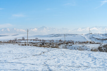 Snowy rural landscapes in Kyrgyzstan in early winter, vast steppes culminate in tall Tian Shan mountains under partly cloudy mostly blue sky.