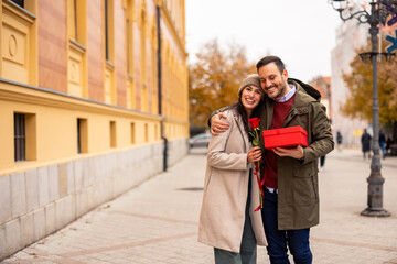 Fototapeta premium Mid adult Caucasian male surprises wife with a present and red rose. They enjoy a romantic day outdoors. She wears a cozy coat and beanie, while he sports a jacket.