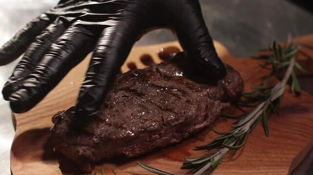 Slow-motion of a hand in black glove serving a grilled steak on wooden board with rosemary