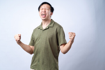 An Asian man in a casual green shirt clenches his fists and grins with excitement, standing against a clean white background. The image conveys joy, success, or celebration.