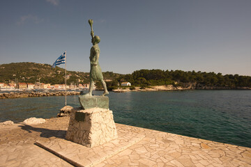 Bronze statue of Georgios Anemogiannis at the entrance to the marina of the village of Gaios on the island of Paxos in Greece with sunset light and the Greek flag