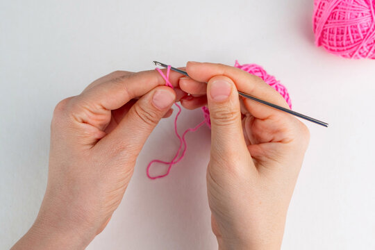 Hand holding thread of pink yarn and a chrocket hook on light background. Tutorial on how to crochet magic ring, step 3