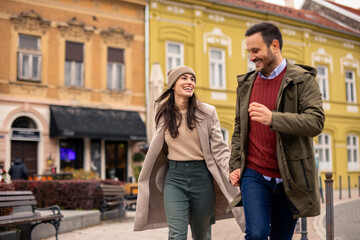 Caucasian couple joyfully running through city streets on Valentine's Day. The man wears a red sweater and green jacket, while the woman sports a cozy beige coat and hat.