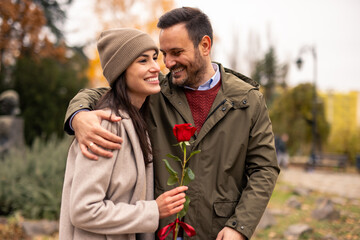 Fototapeta premium Caucasian couple delighting in a public park stroll, woman holding a rose, dressed warmly for Valentine's Day.
