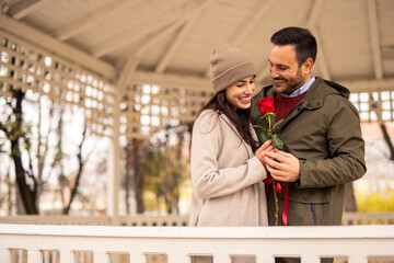Mid adult Caucasian male surprises his girlfriend with a red rose at a pavilion. Both dressed...