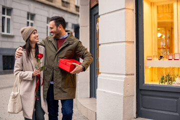 Fototapeta premium A mid adult Caucasian couple walks cheerfully on a city street on Valentine's Day. The man, holding a red gift box, and the woman, with a rose, wear casual winter attire.