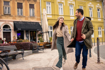 Obraz premium Smiling Caucasian couple enjoys a fun run on the street. Man in a green jacket and red sweater, woman in a beige coat and hat.