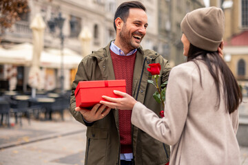 A mid adult Caucasian male surprises his partner with a red rose on a city street. Dressed...