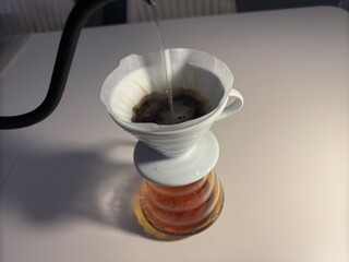 A minimalist pour-over coffee setup featuring a ceramic dripper, paper filter, glass carafe, and a black gooseneck kettle on a white table.