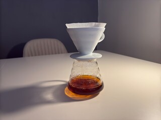 A minimalist pour-over coffee setup featuring a ceramic dripper, paper filter, glass carafe, and a black gooseneck kettle on a white table.