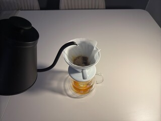 A minimalist pour-over coffee setup featuring a ceramic dripper, paper filter, glass carafe, and a black gooseneck kettle on a white table.