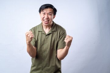 Fototapeta premium An Asian man in a casual green shirt clenches his fists and grins with excitement, standing against a clean white background. The image conveys joy, success, or celebration.
