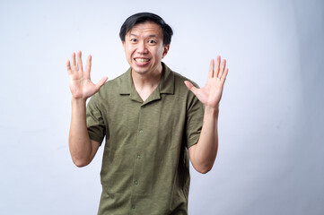 Fototapeta premium An Asian man in a casual green shirt raises his hands and shouts with an exaggerated expression, standing against a clean white background. The image conveys excitement, surprise, or dramatic reaction