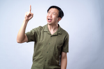 Fototapeta premium An Asian man in a casual green shirt excitedly points upward with one finger, mouth open in expression, standing against a clean white background. The image conveys an idea, realization