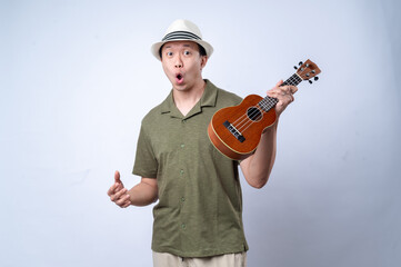An Asian man in a casual green shirt and white hat smiles while playing a ukulele, standing against a clean white background. The image conveys joy, music, or a fun, relaxing activity