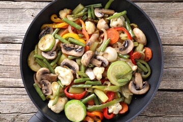 Different vegetables and mushrooms in frying pan on wooden table, top view