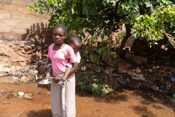 Young African girl standing in a flooded area near her home with her little sister on her back