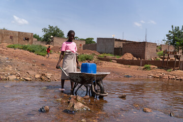 Rural scene of a young African child wading through a river with her heavy wheelbarrow carrying a...