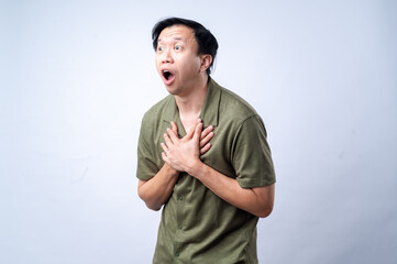 Fototapeta premium An Asian man in a casual green shirt clutches his chest with both hands, expressing pain or discomfort, standing against a clean white background. The image symbolizes chest pain